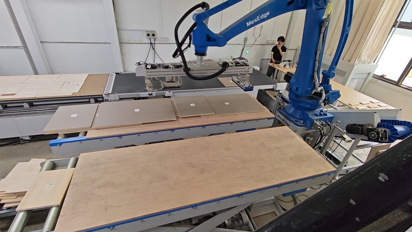 Specifying and Verifying Low-Formaldehyde Plywood on Singapore Projects A robot arm handling cut panel components beside nested parts laid out on the machine tables.